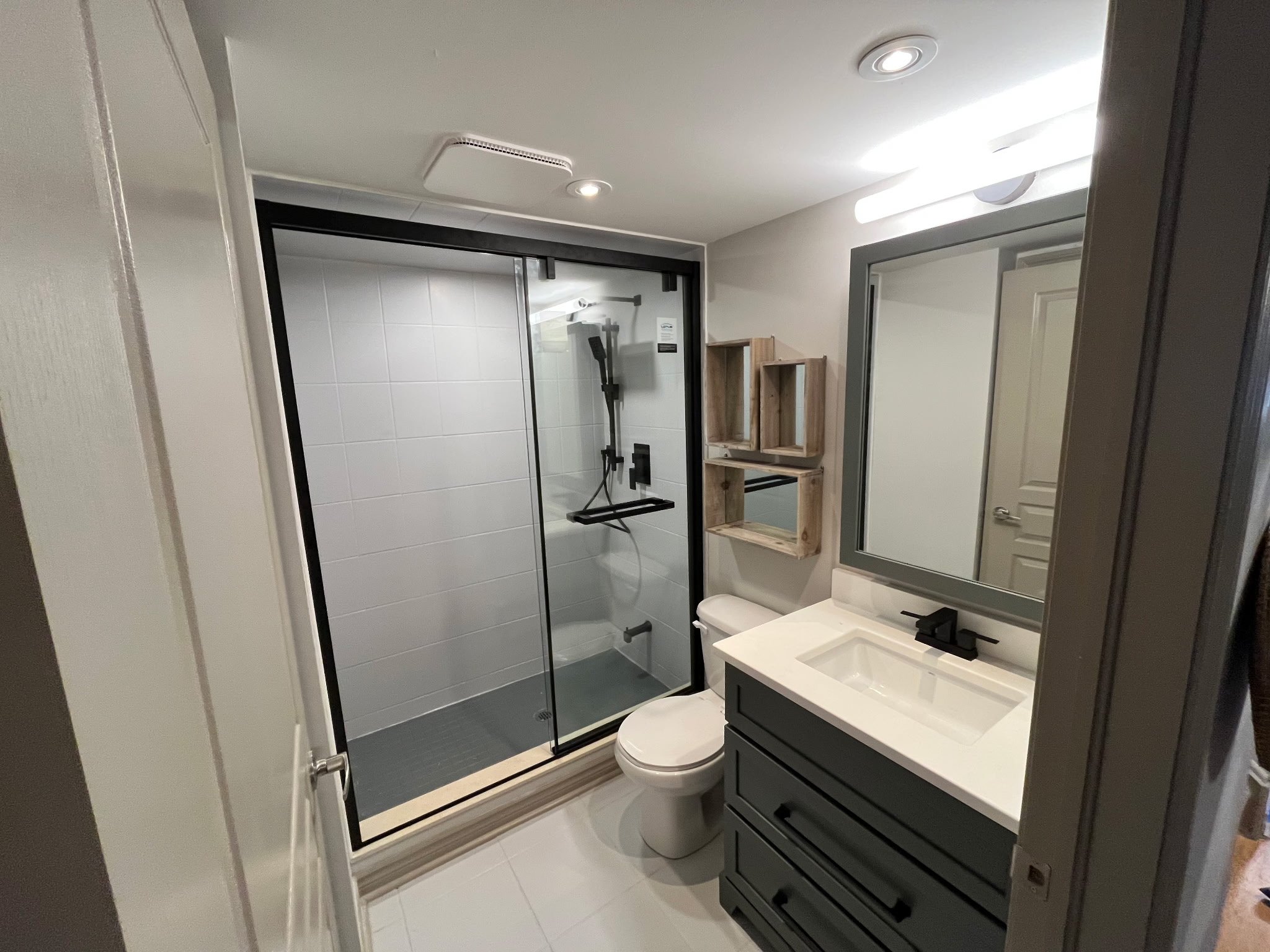 Modern bathroom with a glass-panel shower, toilet, and vanity. The vanity has a large mirror and drawers. Walls are white, and shelving is above the toilet. Ceiling lights are installed.