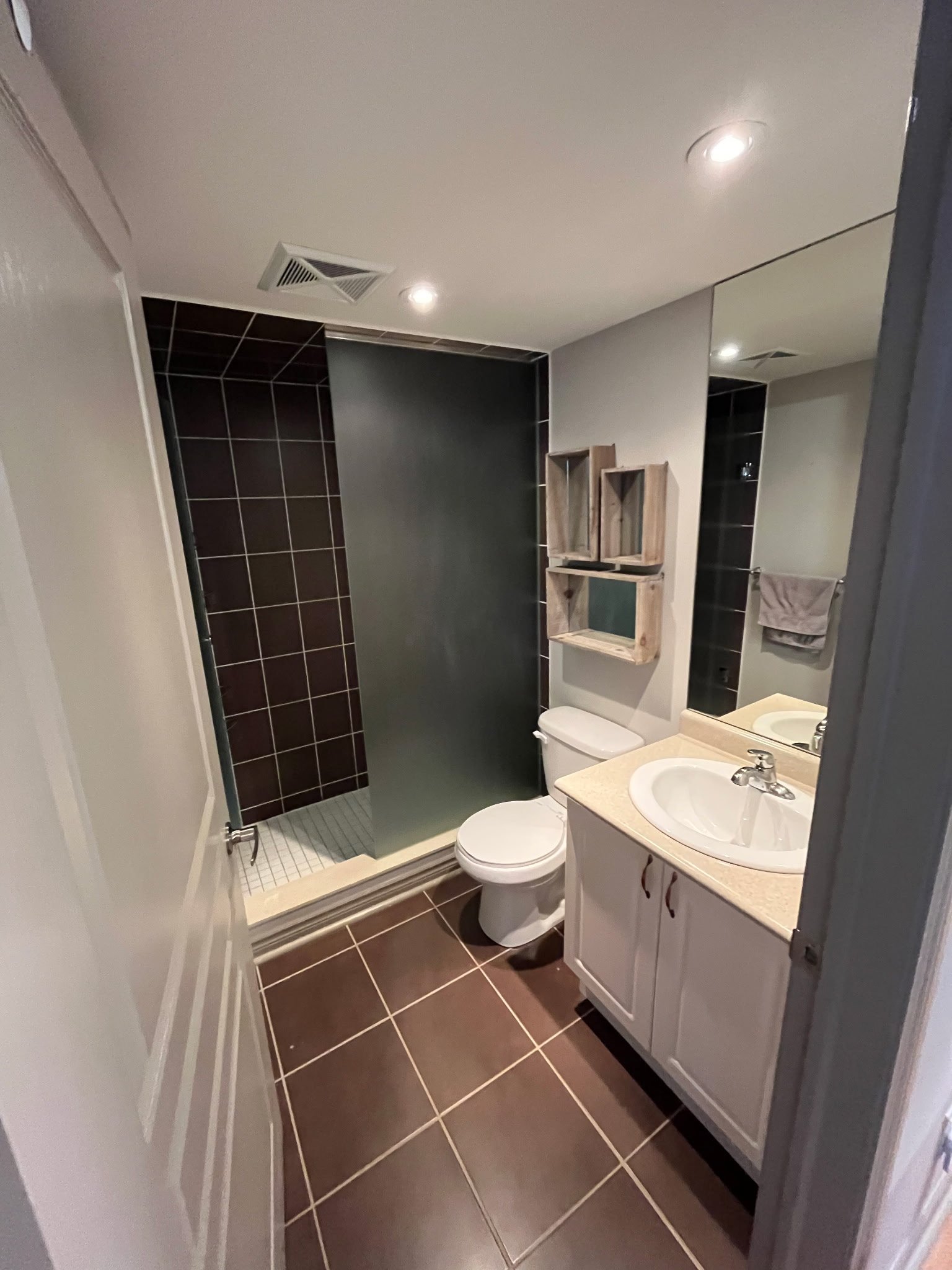 Small bathroom with brown tile flooring, a shower with dark tiles, a white sink with a countertop, a toilet, and a wooden wall shelf above the toilet.