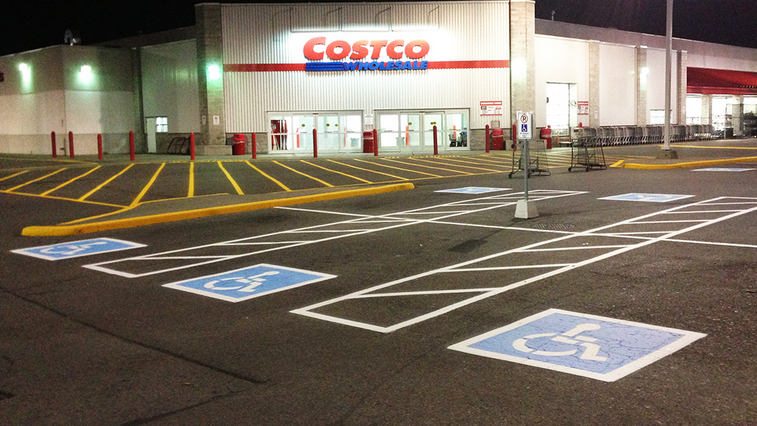 The image shows the entrance of a Costco Wholesale store at night, freshly maintained with Commercial Painting Services in Collingwood. There are several marked handicap parking spaces in the foreground, and the store's brightly lit entrance is visible in the background.