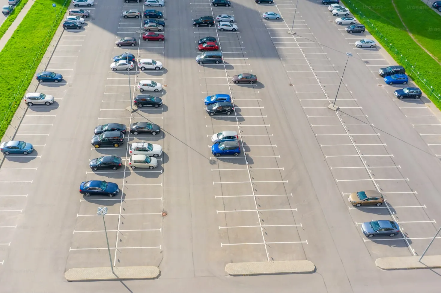 Aerial view of a large parking lot with scattered parked cars and empty spaces, bordered by green grass.